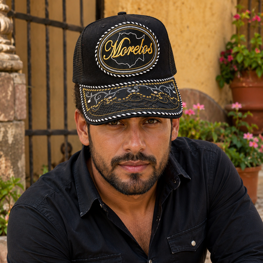 A Mexican model wearing a black Morelos charro-style hat in a warm hacienda courtyard with flowering plants. The embroidered Morelos name and map design are clearly visible on the front.