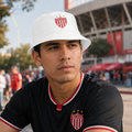 A man wearing a white bucket hat and a black soccer jersey with red and white accents, both featuring the Necaxa logo, photographed outdoors in front of a stadium.