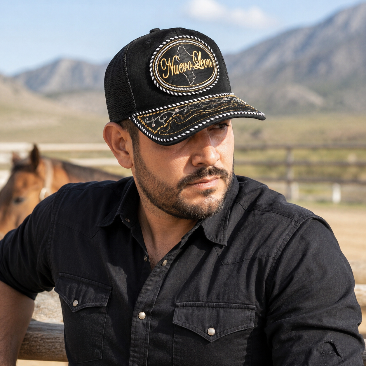 A Mexican model wearing a black Nuevo León charro-style hat at a northern Mexican ranch with mountains in the background. The embroidered Nuevo León name and map design are clearly visible on the front.