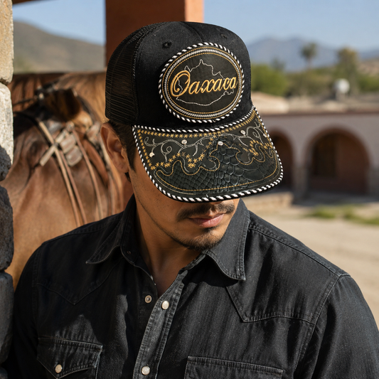 A Mexican man wearing a black Oaxaca adjustable charro-style hat with embroidered gold and white details and a snake-skin design on the front. He is photographed outdoors near a saddle and rustic Mexican-style buildings.