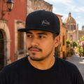A Mexican model wearing the Oaxaca snapback hat in a realistic outdoor lifestyle setting, with the front design clearly visible. The Oaxaca snapback hat design, shape, and colors are shown clearly in a natural casual scene.