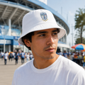 A man wearing a white bucket hat with a blue and gold Pachuca crest, standing in front of a stadium background.