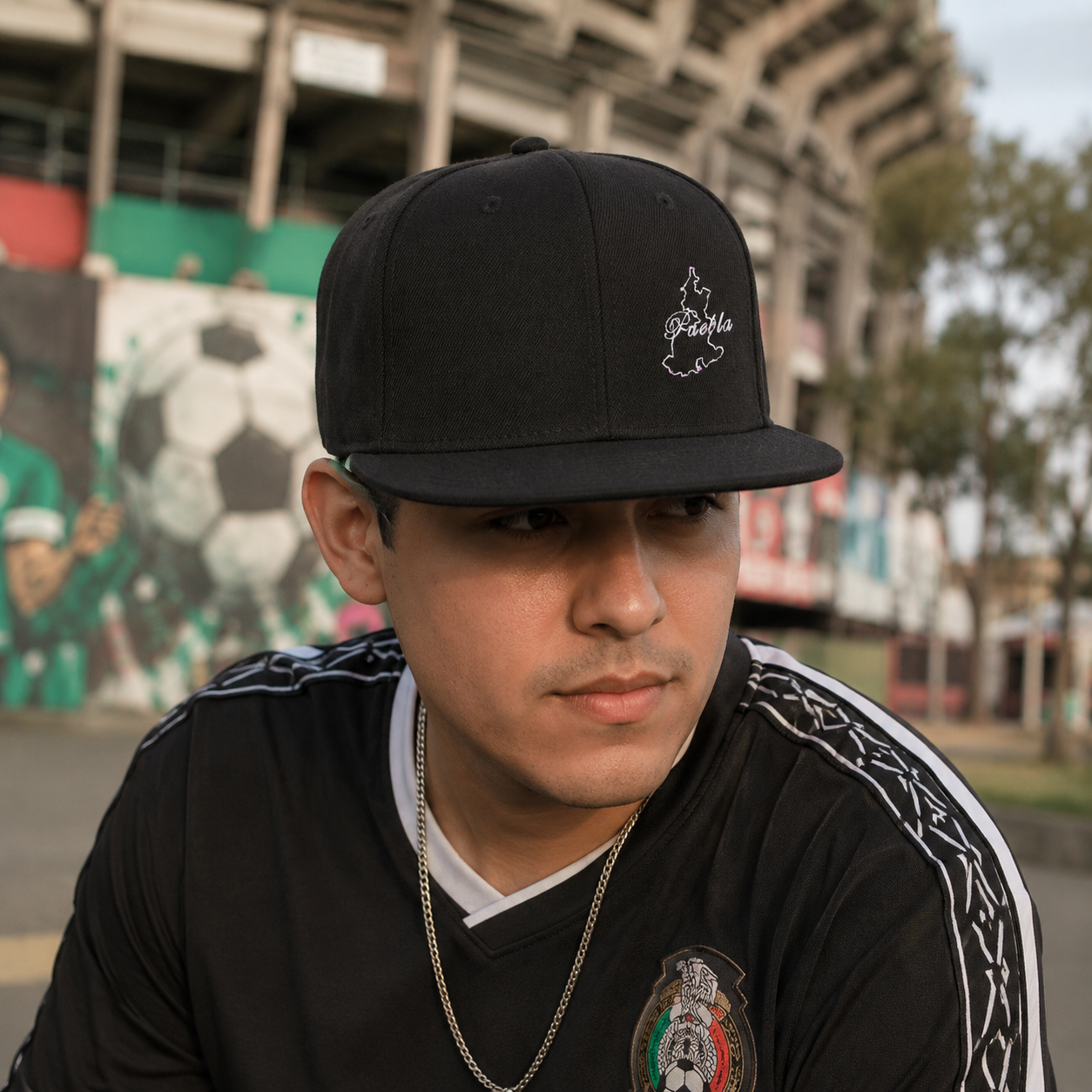 The image shows a man wearing a black snapback hat with a minimalist embroidered outline of the Mexican state of Puebla and the word “Puebla” in cursive white text. He’s dressed in a black soccer jersey featuring Mexico’s national emblem and a subtle pattern along the shoulders. A thin silver chain adds a clean touch to his look.