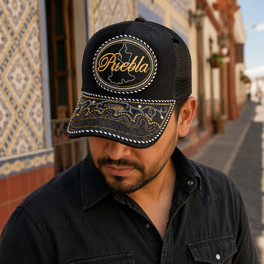 A Mexican model wearing a black Puebla charro-style hat on a colonial street with tiled architecture. The embroidered Puebla name and map design are clearly visible on the front.