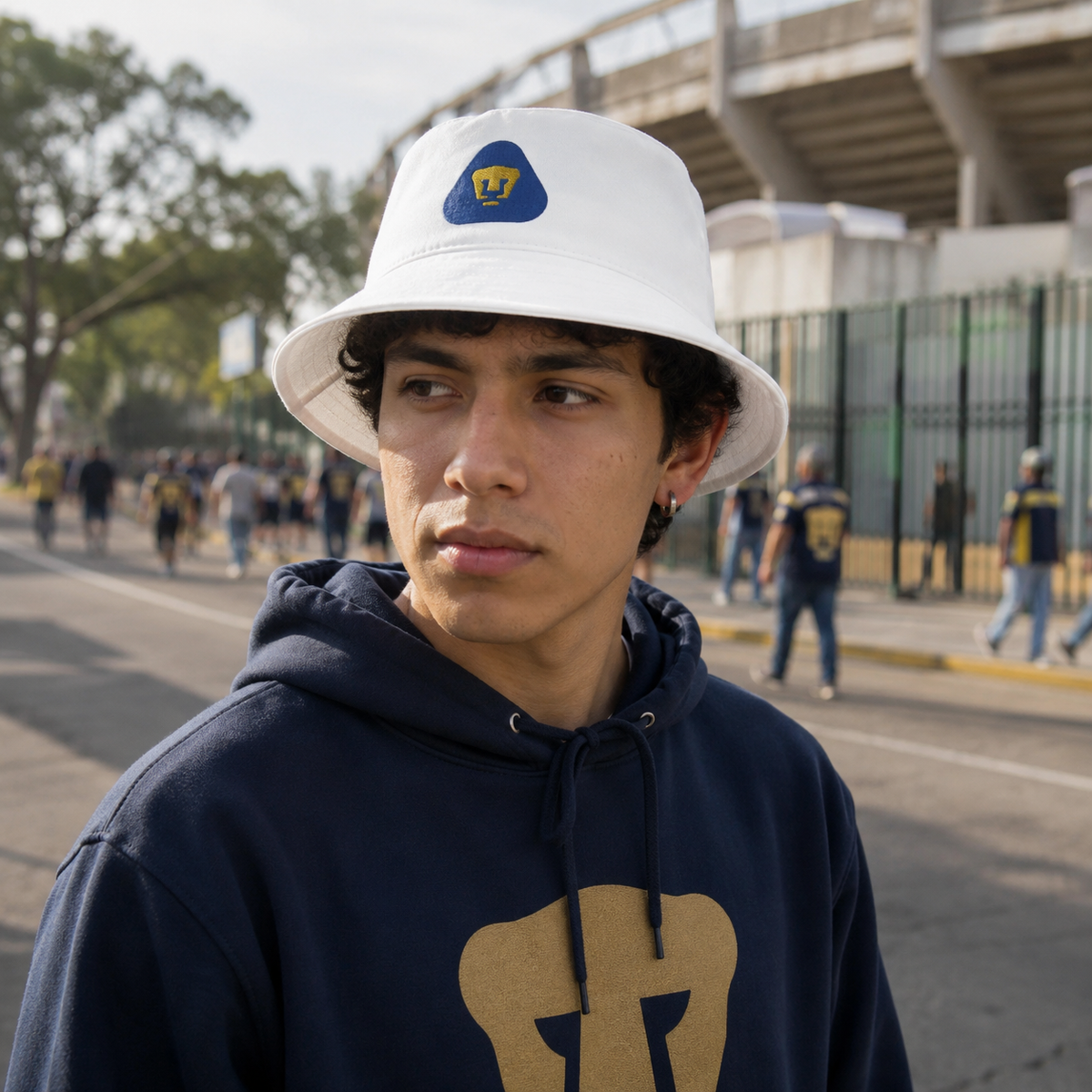 A young man wearing a white bucket hat with a blue and yellow Pumas logo and a navy blue hoodie with a large gold graphic, standing outdoors in a stadium setting.