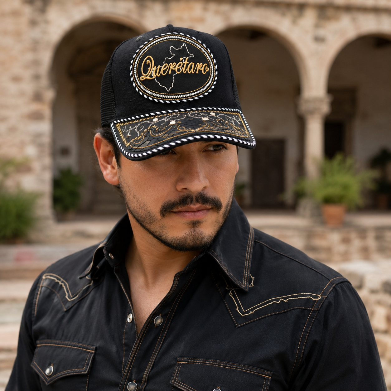 A Mexican model wearing a black Querétaro charro-style hat in a historic courtyard with stone arches. The embroidered Querétaro name and map design are clearly visible on the front.