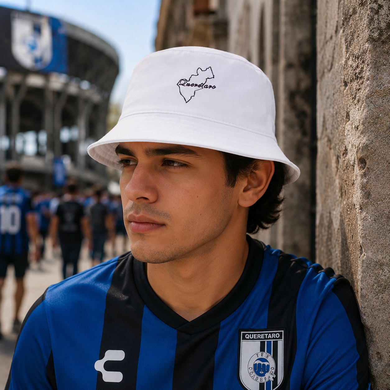 A man wearing a white bucket hat with a black outline of Queretaro and the word 'Queretaro' embroidered on it, leaning against a stone wall. He is wearing a blue and black Queretaro soccer jersey.