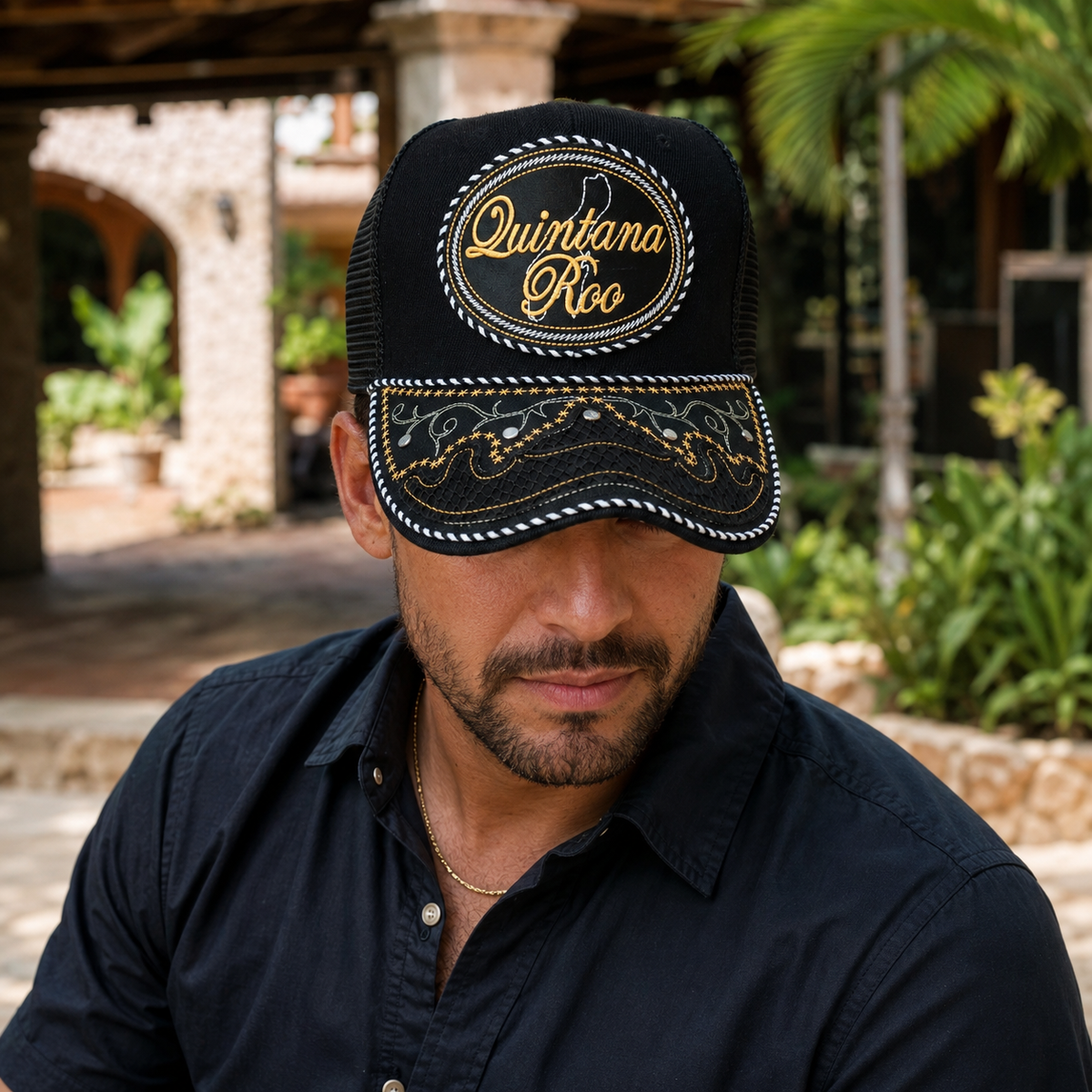 A Mexican model wearing a black Quintana Roo charro-style hat in an outdoor hacienda setting with tropical foliage. The embroidered Quintana Roo name and map design are clearly visible on the front.