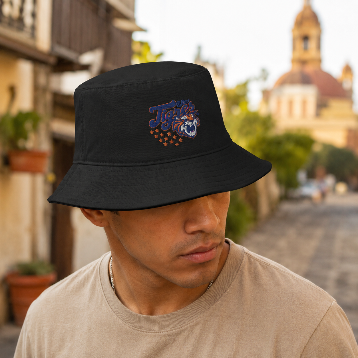 A man wearing a black bucket hat with a colorful embroidered 'Tigres de Quintana Roo' logo, standing outdoors on a blurred street background.