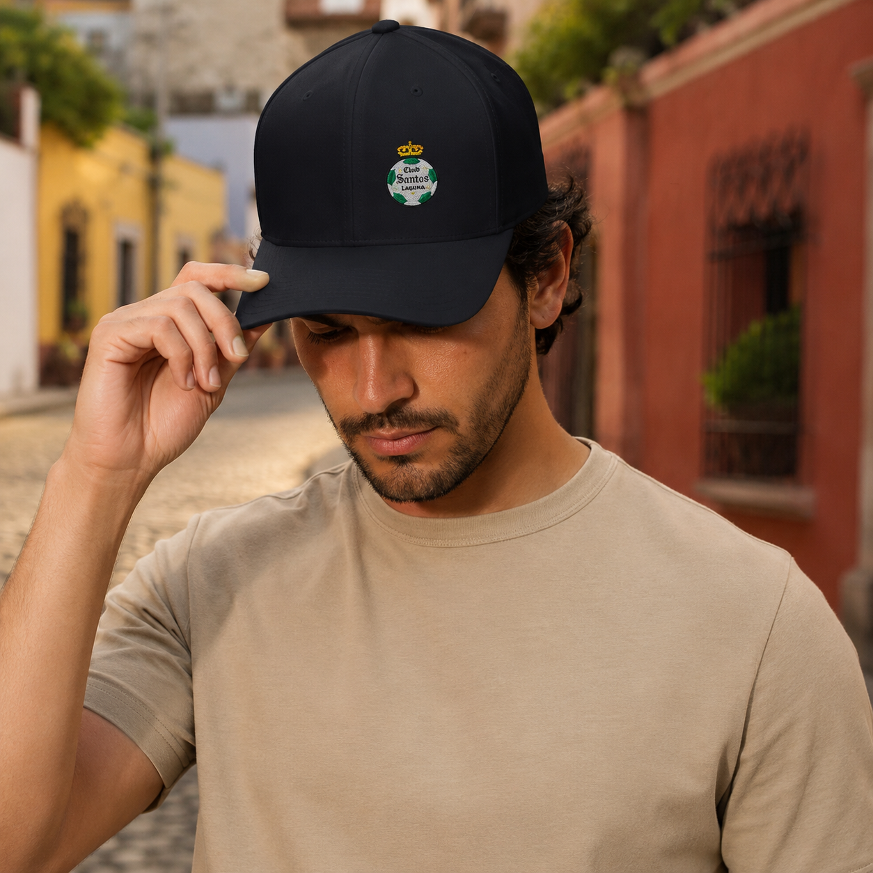 A man wearing a black baseball cap with a small Santos Laguna embroidered crown and shield logo on the front, holding the brim with one hand while wearing a beige t-shirt in an outdoor street setting.
