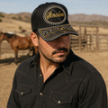A Mexican model wearing a black Sinaloa charro-style hat at a ranch with dry countryside in the background. The embroidered Sinaloa name and map design are clearly visible on the front.