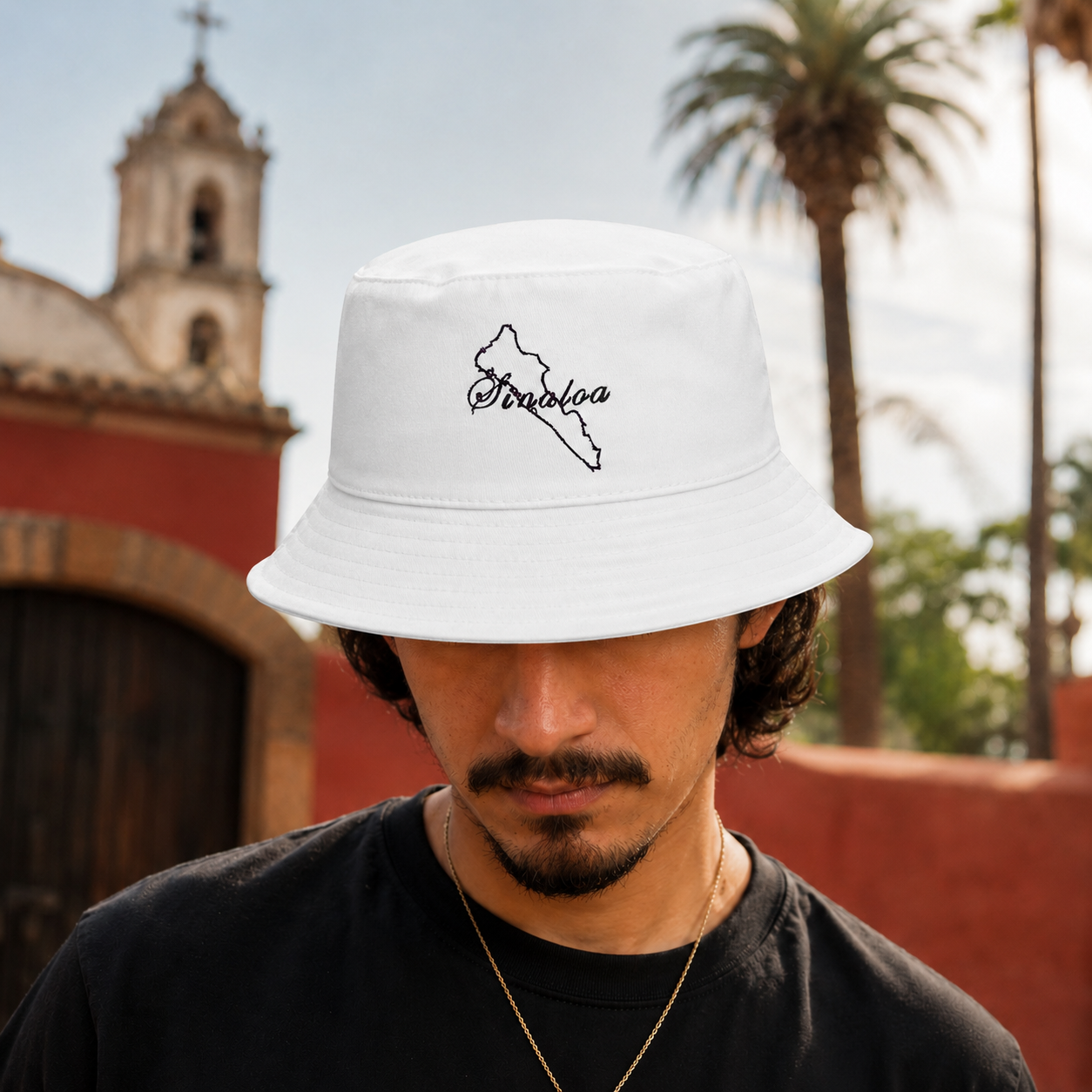 A man wearing a white bucket hat with a black outline of the state of Sinaloa and the word 'Sinaloa' written in script, photographed outdoors in front of a red wall and a church tower.