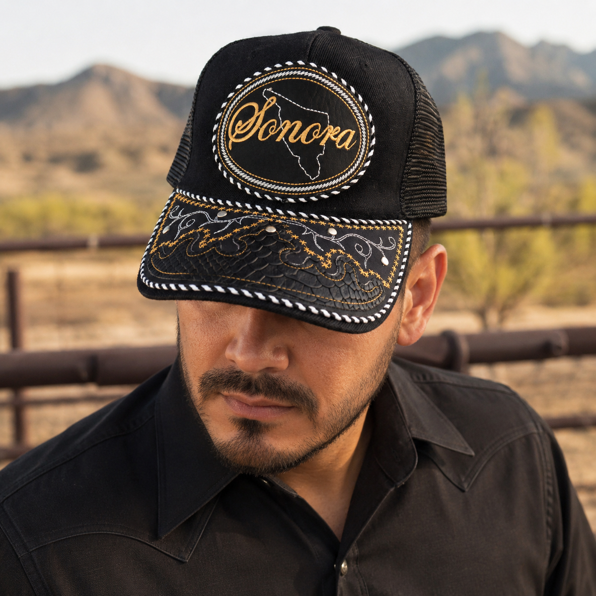 A Mexican model wearing a black Sonora charro-style hat in a desert vaquero setting. The embroidered Sonora name and map design are clearly visible on the front.