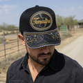 A Mexican model wearing a black Tamaulipas charro-style hat on a northern Mexican ranch road with fencing. The embroidered Tamaulipas name and map design are clearly visible on the front.