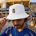 A man wearing a white bucket hat with a blue and yellow Tigres UANL team crest, standing in a stadium crowd. He is wearing a blue and yellow soccer jersey.