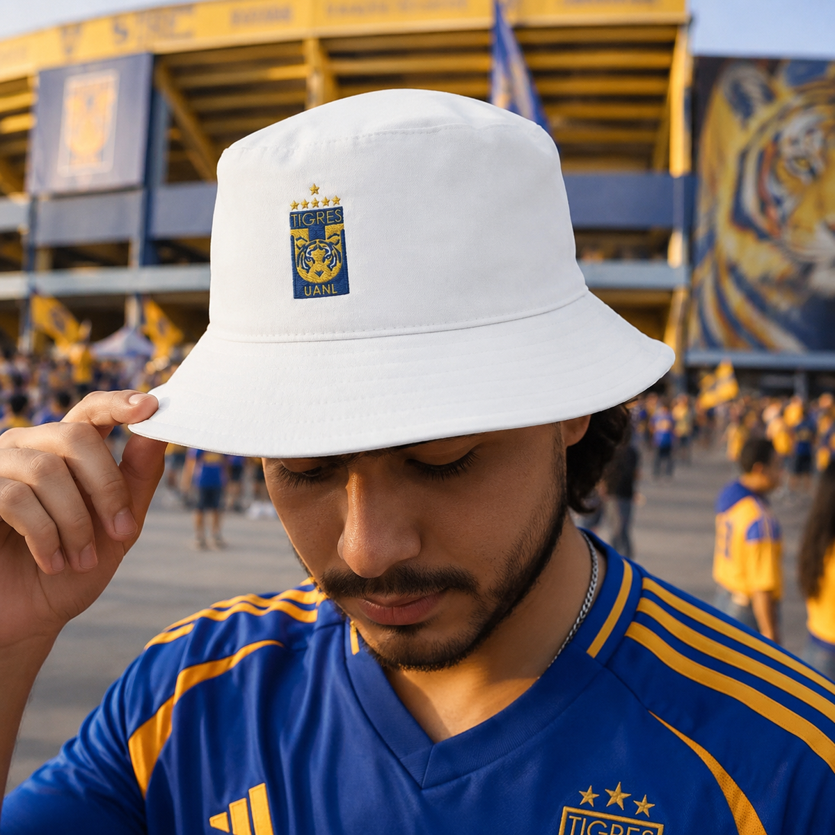A man wearing a white bucket hat with a blue and yellow Tigres UANL team crest, standing in a stadium crowd. He is wearing a blue and yellow soccer jersey.