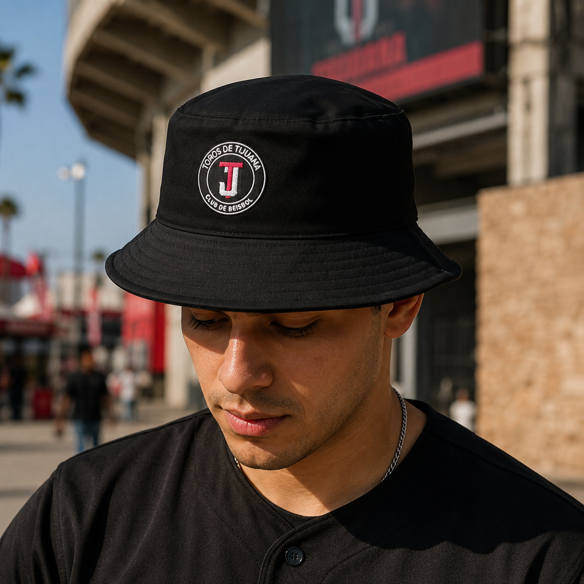 A man wearing a black bucket hat with a Toros de Tijuana embroidered logo on the front, photographed outdoors in front of a stadium or arena.