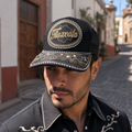 A Mexican model wearing a black Tlaxcala charro-style hat on a historic village street. The embroidered Tlaxcala name and map design are clearly visible on the front.