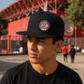 A man wearing a black baseball cap with a Deportivo Toluca Mexican national team logo, photographed outdoors in front of a red stadium.