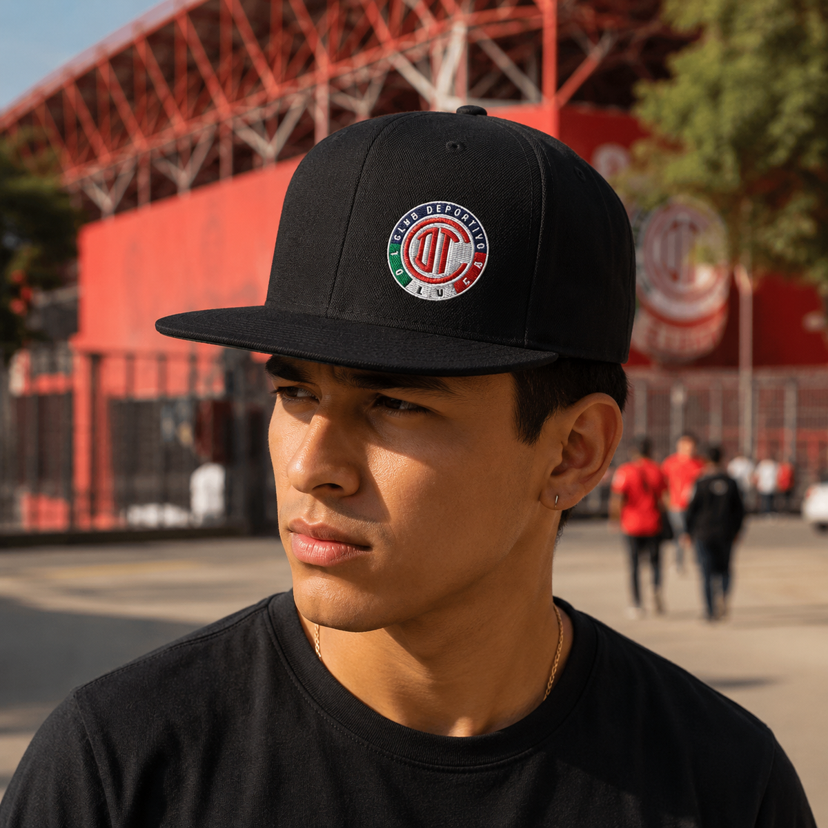 A man wearing a black baseball cap with a Deportivo Toluca Mexican national team logo, photographed outdoors in front of a red stadium.