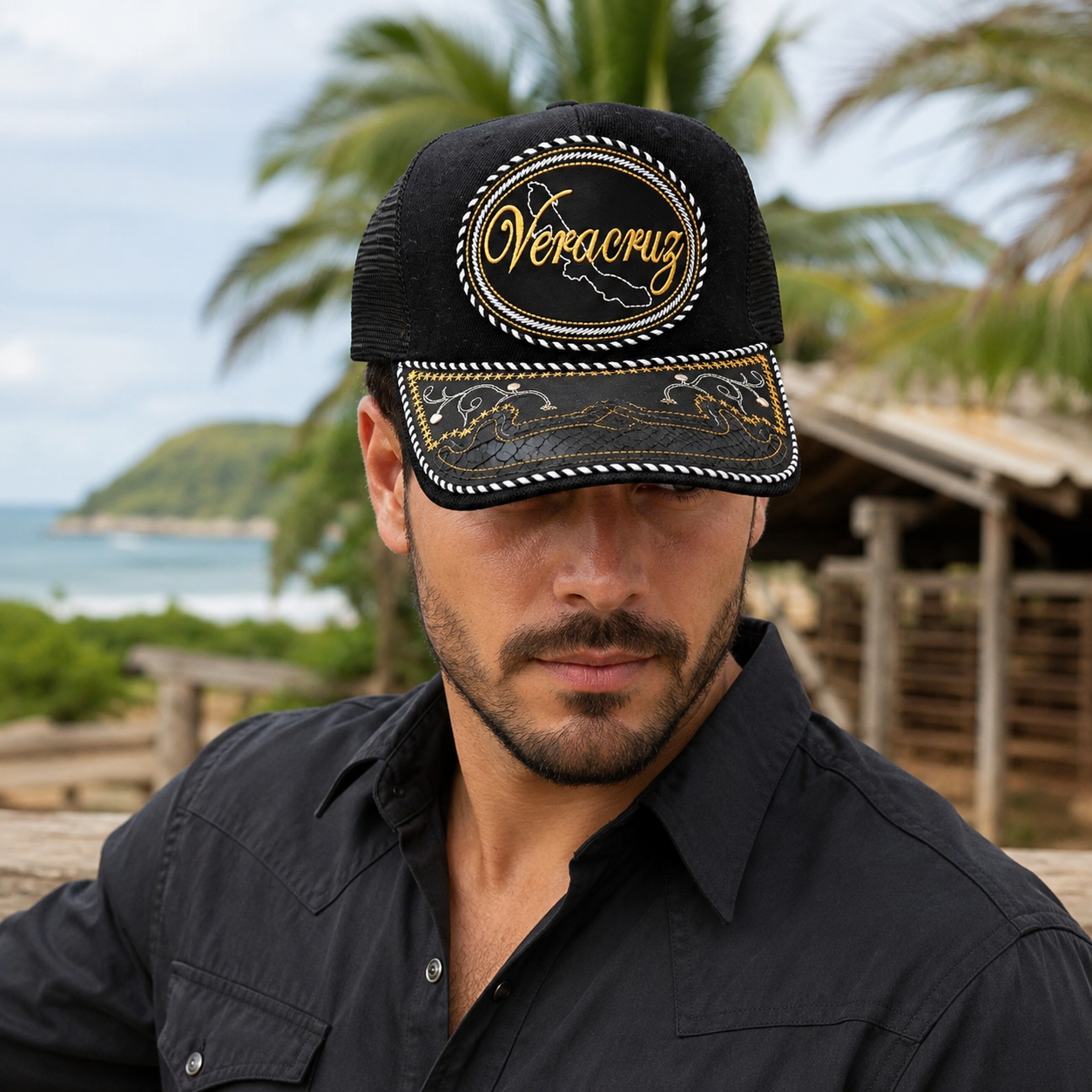 A Mexican model wearing a black Veracruz charro-style hat in a coastal Mexican ranch setting. The embroidered Veracruz name and map design are clearly visible on the front.