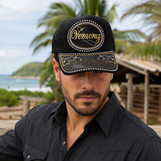 A Mexican model wearing a black Veracruz charro-style hat in a coastal Mexican ranch setting. The embroidered Veracruz name and map design are clearly visible on the front.