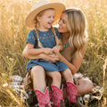 A woman and a young girl sitting together in a golden grassy field at sunset. The girl wears a straw cowboy hat, a blue dress with floral embroidery, and bright pink cowboy boots with white swirl patterns.