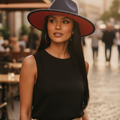 A woman wearing a black sleeveless top, beige trousers, and a wide-brimmed navy hat with a red underside stands on a cobblestone street in an outdoor urban setting.