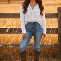 A woman stands outdoors in a sunlit field in front of a wooden fence, wearing a white long-sleeve button-up shirt, blue jeans, and brown cowboy boots.
