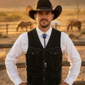 A man wearing a black western vest over a white button-up shirt with a blue bolo tie, standing outdoors in a ranch setting with mountains and horses in the background.