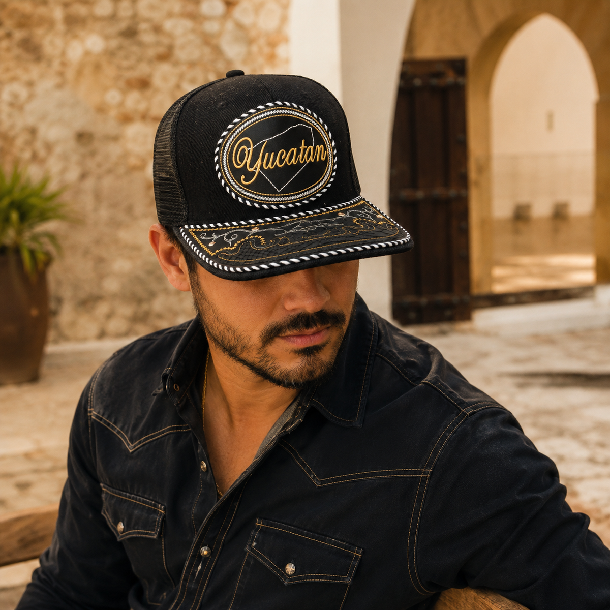 A Mexican model wearing a black Yucatán charro-style hat in a hacienda courtyard with limestone walls. The embroidered Yucatán name and map design are clearly visible on the front.