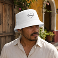 A man wearing a white bucket hat with 'Yucatan' embroidered in black script, set against a warm, sunlit outdoor background with a wooden door and yellow walls.