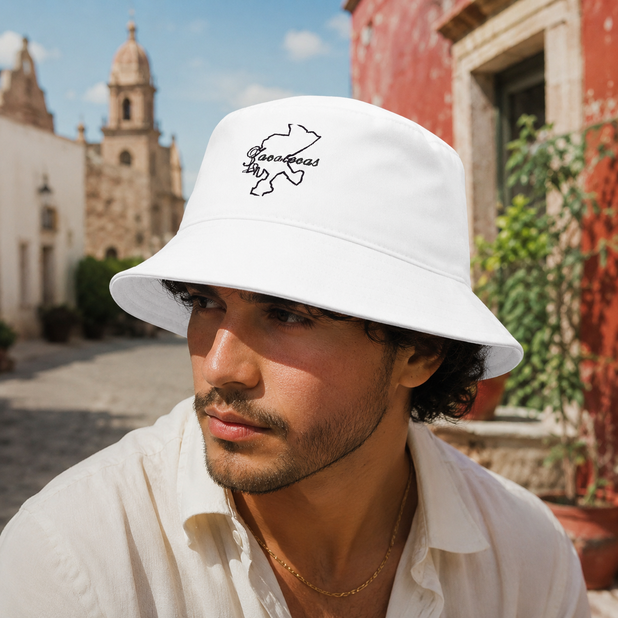 A man wearing a white bucket hat with a black outline logo that reads 'Zacatecas' and a white shirt, photographed outdoors in a sunlit street with historic buildings in the background.