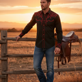 A man stands outdoors at sunset wearing a black long-sleeve western-style button-up shirt with red embroidered detailing on the chest and cuffs, paired with blue jeans and dark boots. He is leaning on a wooden fence next to a brown saddle in a dry field.